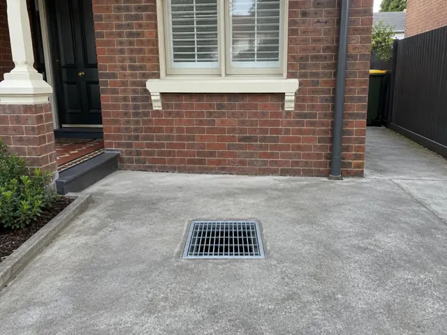 Clean outdoor drain grate set in a concrete driveway beside a brick house in Lewisham